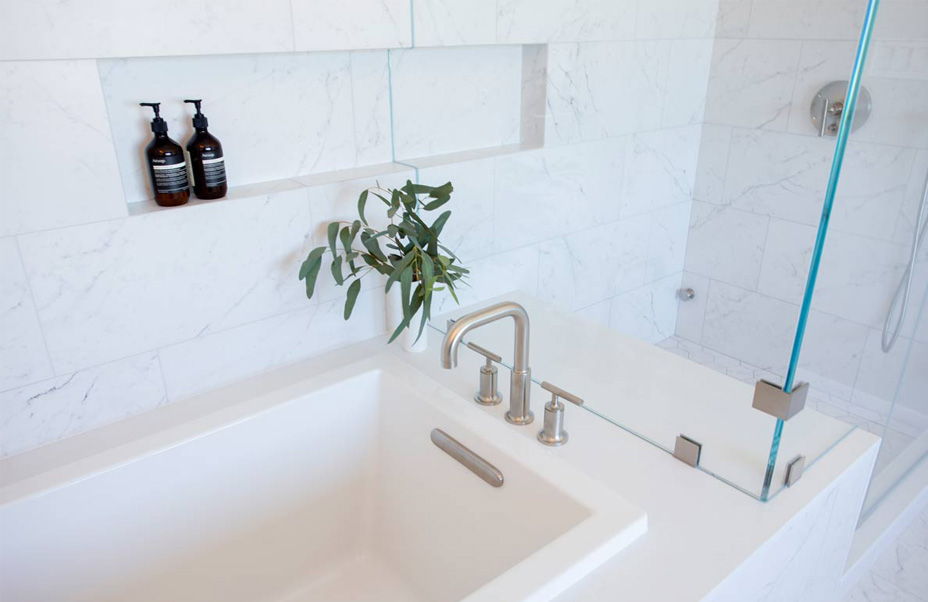 A modern bathroom featuring a white bathtub next to a glass shower partition. The walls are tiled in white marble. A recessed shelf above the tub holds two dark bottles and a plant branch. A sleek silver faucet is installed on the tub edge