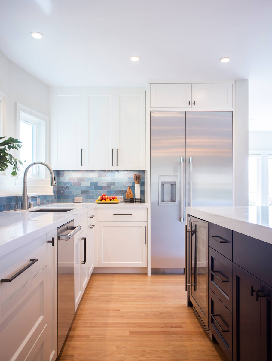 A modern kitchen remodel in San Francisco features white cabinets, stainless steel appliances, and a blue tile backsplash. The large refrigerator, sink under the window, and an island with dark drawers complement the light wood flooring, enhancing the bright and airy feel