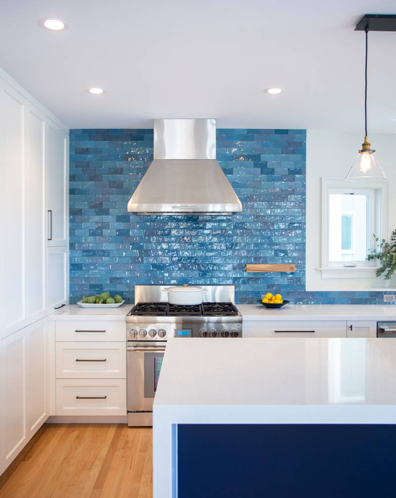 Modern kitchen with a stainless steel range and hood, surrounded by blue textured tiles. White cabinets and countertops contrast with the blue accents. A pendant light hangs above, with lemons and limes adding a touch of color