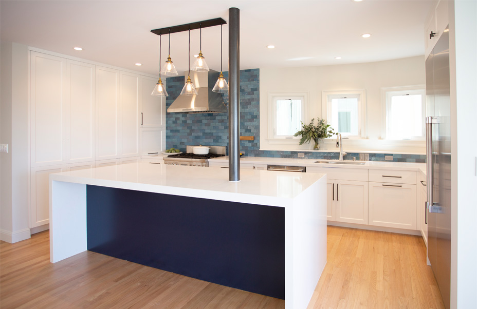 In a recent San Francisco remodel, this modern kitchen features white cabinetry, a stainless steel stove and hood, and a blue tile backsplash. A white island countertop sits in the foreground, illuminated by a pendant light above. Lemons and limes add a vibrant touch to the counter.