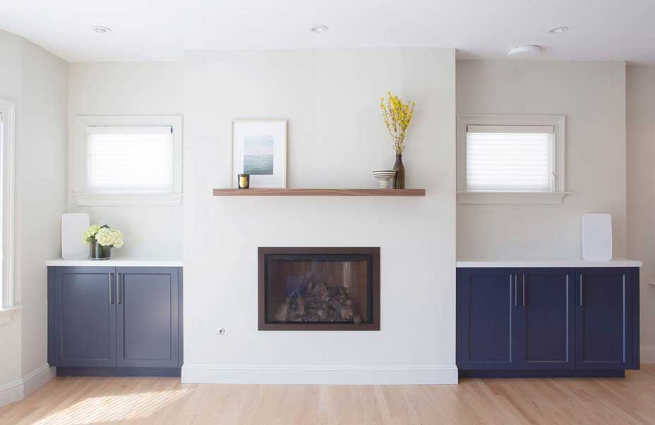 A modern living room with a central fireplace. The walls are light-colored, and the floor is wooden. Blue cabinets flank the fireplace, and a wooden shelf above it holds a framed picture and a vase with yellow flowers.