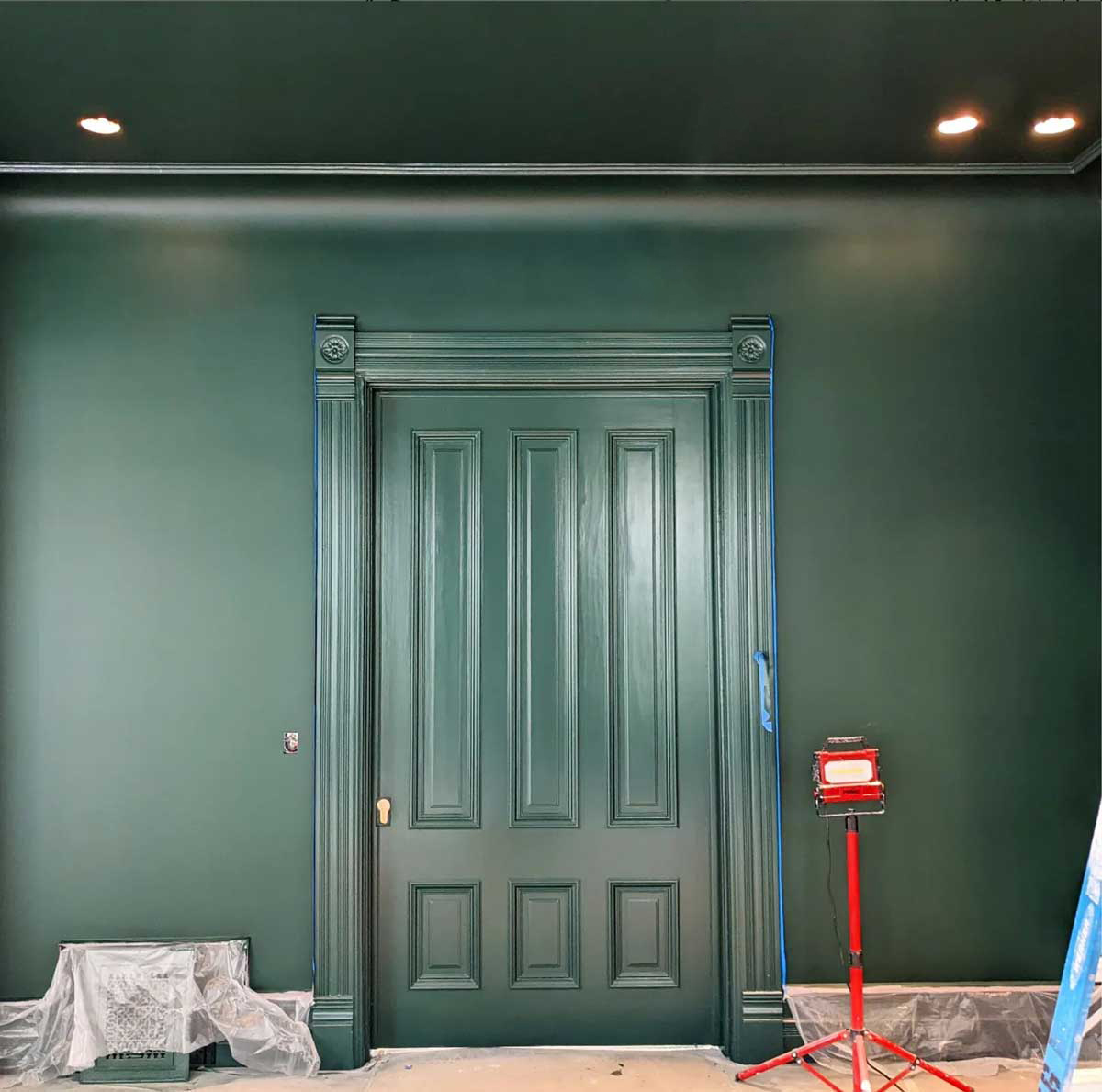 A room with dark green walls and a matching door. Trim and panels are also painted green. A red construction light is placed on the floor to the right, and plastic covers protect the floor and small parts of the walls