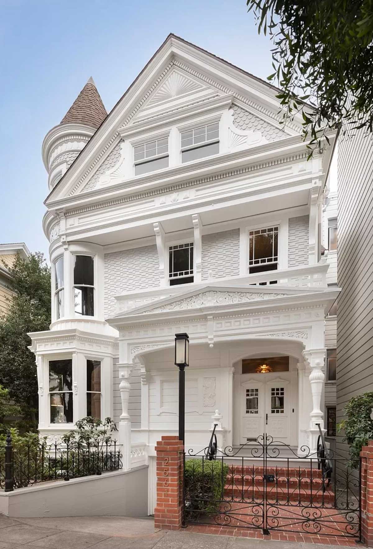 A large, ornate Victorian-style house painted white with intricate woodwork and a steep roof. It features a turret on the left, tall windows, and a brick staircase leading to a decorative wrought iron gate. Trees frame the house.