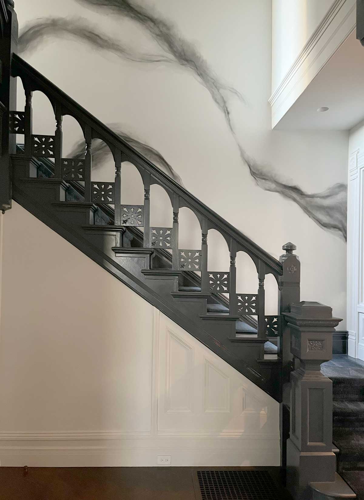 A black wooden staircase with ornate railing details, featuring arches and intricate patterns, leads upward alongside a white wall adorned with a swirling, dark gray design. The flooring at the bottom is dark and contrasts with the lighter wall.